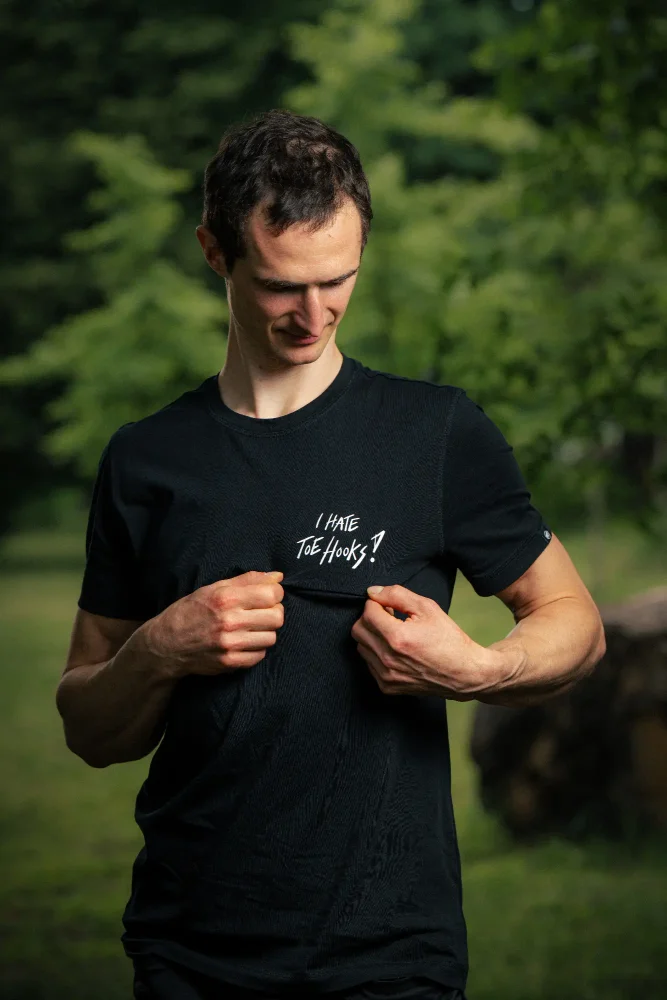 Adam Ondra showing the "I hate toehooks" sign on his black T-shirt